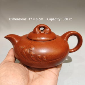 Puede incluir: Una tetera de arcilla de color marrón rojizo con asa curva, pico corto y tapa con asa en forma de bucle. El cuerpo presenta patrones de olas en relieve. El texto indica unas dimensiones de 17 x 8 cm y una capacidad de 380 cc.