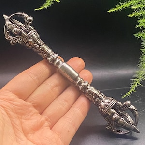 May include: An intricately designed silver vajra, a ritual object with detailed floral and geometric patterns. The vajra is held in a hand, highlighting its symmetrical form. The object has a handle and two opposing prongs.