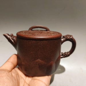 May include: A dark brown clay teapot with a textured, matte finish. The lid features intricate embossed patterns. The spout and handle are sculpted into dragon-like shapes, with the handle showing detailed scales and a curved, ornate design.