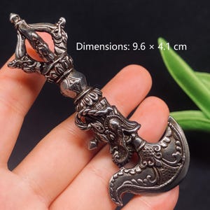 May include: A silver-toned metal ritual axe with intricate embossed dragon motifs and a vajra-style handle. The object features detailed scrollwork and measures 9.6 x 4.1 cm. The design includes a curved blade and a multi-pronged top section.