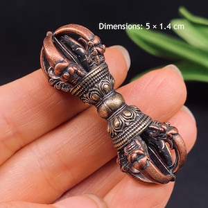 May include: A copper-toned metal vajra ritual object featuring intricate relief carvings of mythical creatures and lotus petal patterns. The object measures 5 x 1.4 cm as indicated by the text in the image. The design is symmetrical with curved prongs at both ends and a central grip.