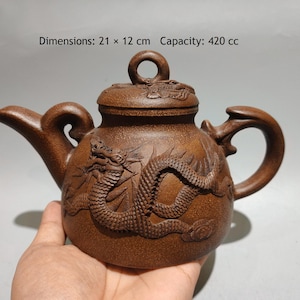 Puede incluir: Una tetera de arcilla marrón con un diseño de dragón en relieve en el cuerpo y la tapa, con un asa circular en la parte superior. La tetera mide 21 x 12 cm con una capacidad de 420 cc. El texto Dimensions: 21 x 12 cm Capacity: 420 cc es visible.