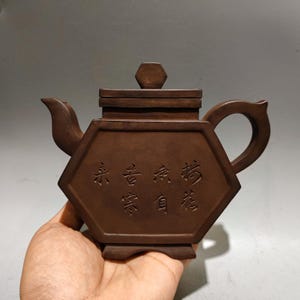 May include: A hexagonal brown clay teapot featuring a flat lid with a hexagonal knob, a curved spout, and a handle. The front side of the teapot is embossed with two vertical columns of Chinese characters, showcasing a matte, earthy finish.