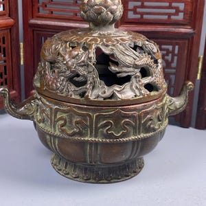 Op de afbeelding: Bronzen wierookbrander met ingewikkelde reliëfs van draken en bloemen. Het deksel heeft een lotusbloem-knop. De brander heeft twee gebogen handvatten en een decoratieve basis. Het ontwerp is sierlijk en gedetailleerd.