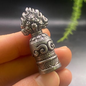 Può includere: Un pendente a forma di campana color argento, ricco di dettagli. La parte superiore presenta un motivo floreale, mentre il corpo mostra un'intricata lavorazione a volute. Il pendente è tenuto in mano, evidenziando le sue dimensioni. Un gioiello unico.