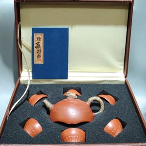 Peut inclure: Une théière en argile brune avec un animal sculpté sur le couvercle et une anse texturée, accompagnée de cinq petites tasses assorties, le tout dans une boîte de présentation brune doublée de mousse avec un livret bleu portant le texte 珍藏證書.