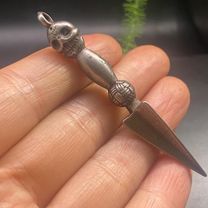 May include: A silver-toned pendant in the shape of a small dagger. The handle features a skull at the top, a woven knot design in the middle, and a pointed blade. The pendant is held in a hand.