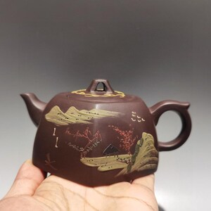 May include: A dark brown hexagonal clay teapot featuring a lid with a small handle. The body is decorated with a painted landscape scene in muted green, red, and yellow tones, depicting mountains, trees, and small structures on its flat sides.