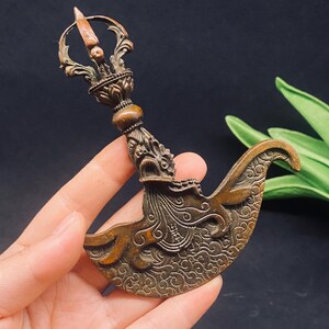 Könnte beinhalten: Ein bronzefarbenes rituelles Hackmesser, bekannt als Kartika, mit einer geschwungenen Klinge mit komplizierten Wirbelmustern und einem Griff, der mit einem Vajra-Endstück verziert ist. Die Metalloberfläche zeigt eine verwitterte Patina.