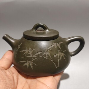 May include: A dark brown clay teapot featuring a rounded body, a curved spout, and a handle. The exterior surface is decorated with an engraved bamboo motif and includes Chinese characters etched into the side. It has a matching lid with a loop.
