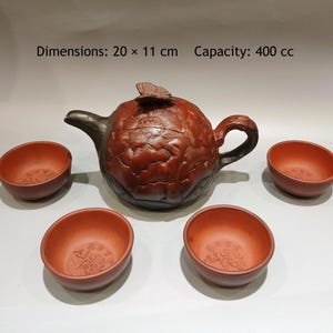 May include: A reddish-brown clay teapot with a sculpted floral pattern and a butterfly lid finial, accompanied by three matching small tea bowls. The teapot measures 20 x 11 cm with a 400 cc capacity. The set features dark accents on the spout and handle.