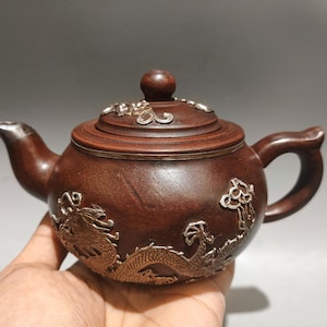 May include: A dark brown clay teapot featuring silver-colored metal dragon relief ornamentation on the body and lid. The teapot has a curved spout with a silver-colored metal tip, a rounded handle, and a matching lid with a central knob.