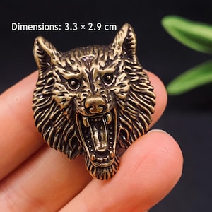 Könnte beinhalten: Ein bronzefarbenes Metall-Emblem in Form eines Wolfskopfes mit knurrendem Ausdruck, sichtbaren Zähnen und texturierten Fell-Details. Der aufgedruckte Text Dimensions: 3.3 x 2.9 cm gibt die physische Größe des Objekts an.