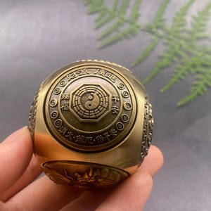 May include: A brass-toned metal sphere featuring an intricate central octagonal medallion with a yin-yang symbol. The surface is engraved with Chinese characters, circular coin motifs, and a relief of a dragon along the lower curved edge.