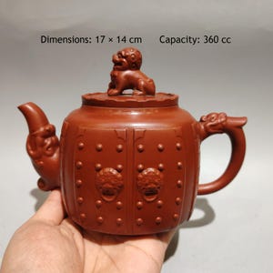 Può includere: Una teiera in argilla color marrone rossiccio con un coperchio sormontato da una figura di leone scolpita. Il corpo presenta un design a forma di porta con borchie in rilievo e battenti a testa di leone. Dimensioni 17 x 14 cm, capacità 360 cc.