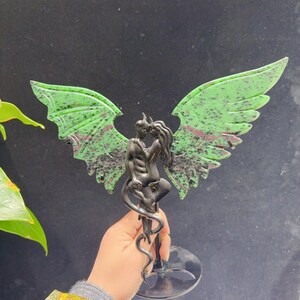 May include: A sculpture featuring two figures embracing, with green and black speckled wings. The figures are dark, and the wings are a vibrant green with red accents. The sculpture is mounted on a clear, circular base.