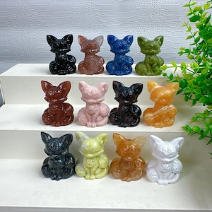 May include: A collection of twelve small fox figurines carved from various colored gemstones, arranged in three rows on a white tiered display. The foxes feature pointed ears, detailed facial features, and curled tails in black, pink, blue, green, and orange.