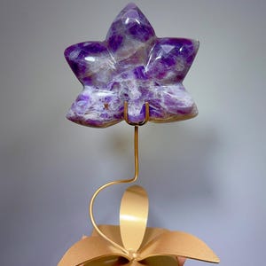 May include: A star-shaped purple and white amethyst crystal carving mounted on a thin gold-colored metal wire stem. The stem is attached to a gold-colored metal base shaped like a flat flower with four petals, providing a stable support.