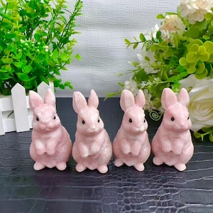 May include: Four pink bunny figurines. Each bunny is in a seated position with upright ears and small, dark eyes. The bunnies are made of a smooth, possibly stone-like material. They are arranged in a row against a dark surface, with greenery in the background.