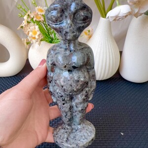 May include: A gray and black stone alien figurine with large eyes and a rounded head. The alien figure is standing on a round base. The figurine is held in a person's hand. The background includes white vases and flowers.