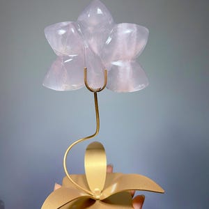 May include: A carved pale pink rose quartz flower sculpture mounted on a thin, curved gold-toned metal stem. The stem is attached to a flat, gold-toned metal base shaped like a stylized four-petaled flower, providing a stable foundation.