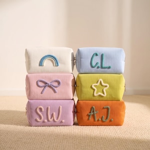 Puede incluir: Una pila de seis estuches rectangulares de pana en varios colores. Arriba a la izquierda, blanco con un arcoíris azul; a la derecha, azul claro con letras verdes C.L.; en el medio, rosa con un lazo morado y amarillo con una estrella blanca; abajo, lavanda con letras rosas S.W. y naranja con letras verdes A.J.