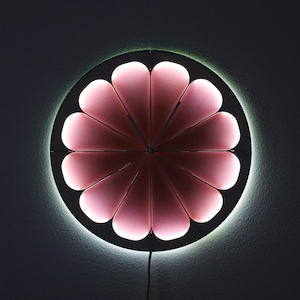 May include: A modern wall clock with a unique design. The clock features a circular black frame with pink, petal-shaped segments that illuminate. A soft white light outlines the clock's edge, and the clock hands are visible.