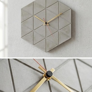 Könnte beinhalten: Eine geometrische Wanduhr mit sechseckiger Form, die aus grauen Dreiecksfliesen besteht. Die Uhr hat goldfarbene Zeiger und einen roten Sekundenzeiger. Die Uhr ist an einer hellgrauen Wand befestigt.