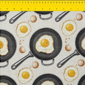 May include: Fabric with a repeating pattern of fried eggs in black skillets, with eggshells scattered around. The eggs have bright yellow yolks and white egg whites. The skillets have silver handles, all on a light beige background.