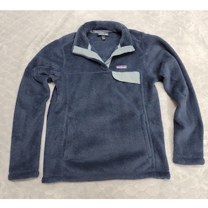 May include: A navy blue fleece pullover with a light blue collar and pocket detail. The pullover has a snap closure at the neck and a small logo patch on the chest. The long-sleeved top is made of a soft, textured material.