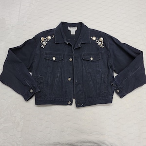 May include: Dark blue denim jacket with silver buttons and pearl and rhinestone embellishments on the shoulders. The jacket has a classic design with two chest pockets and two side pockets.