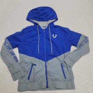 May include: Blue and gray hooded jacket with a full zipper closure. The jacket features a blue hood and a white drawstring. The sleeves are gray with blue text. A white logo is on the chest.