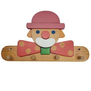 May include: Wooden clown-shaped coat rack with a pink hat and bow tie with red polka dots. The clown has blue eyes, a red nose, and a white mouth. The rack has five wooden pegs for hanging items.