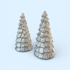 Peut inclure: Deux figurines argent&eacute;es en forme de sapin de No&euml;l textur&eacute;s. Les arbres ont un design en couches, chaque couche ressemblant &agrave; des branches. Les figurines sont pos&eacute;es sur un fond bleu clair, cr&eacute;ant un th&egrave;me festif et d&eacute;coratif.