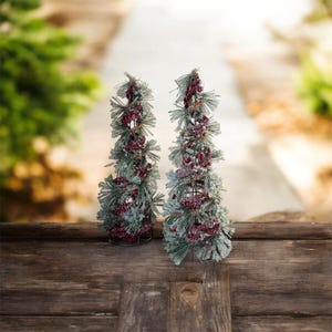 Peut inclure: Deux petits arbres de No&euml;l d&eacute;coratifs. Chaque arbre est compos&eacute; d'aiguilles de pin vertes givr&eacute;es et orn&eacute; de baies rouges. Les arbres sont pos&eacute;s sur une surface en bois sombre avec un arri&egrave;re-plan flou d'une all&eacute;e et de verdure.