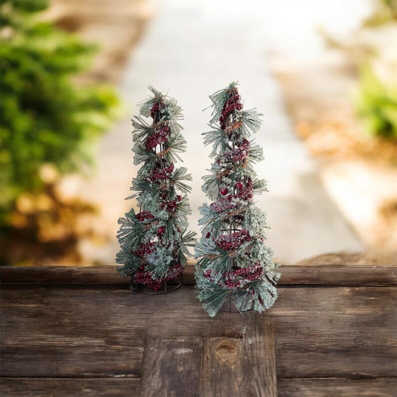 Peut inclure: Deux petits arbres de No&euml;l d&eacute;coratifs. Chaque arbre est compos&eacute; d'aiguilles de pin vertes givr&eacute;es et orn&eacute; de baies rouges. Les arbres sont pos&eacute;s sur une surface en bois sombre avec un arri&egrave;re-plan flou d'une all&eacute;e et de verdure.
