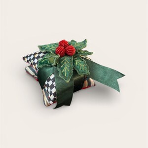 Peut inclure: Un cadeau de f&ecirc;te d&eacute;coratif, compos&eacute; d'une pile de carr&eacute;s de tissu &agrave; motifs, surmont&eacute; d'une d&eacute;coration de houx vert et de baies rouges. Un ruban vert fonc&eacute; est nou&eacute; autour du cadeau. Les carr&eacute;s de tissu pr&eacute;sentent un motif &agrave; carreaux noir et blanc.