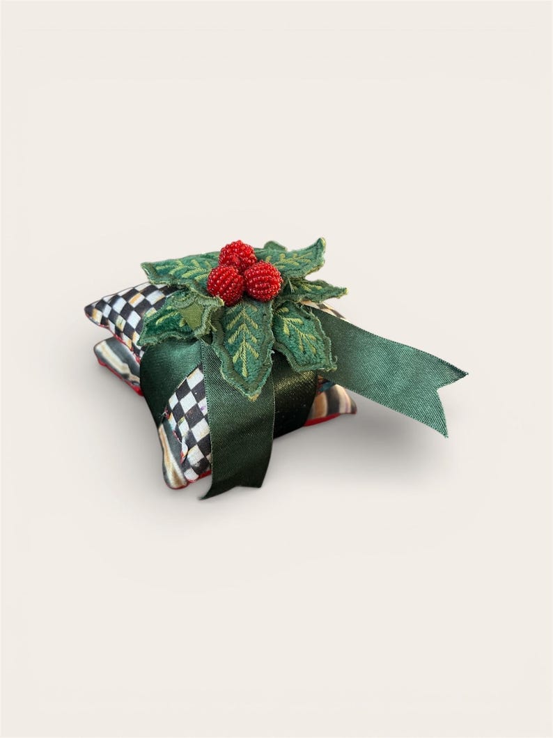 Peut inclure: Un cadeau de f&ecirc;te d&eacute;coratif, compos&eacute; d'une pile de carr&eacute;s de tissu &agrave; motifs, surmont&eacute; d'une d&eacute;coration de houx vert et de baies rouges. Un ruban vert fonc&eacute; est nou&eacute; autour du cadeau. Les carr&eacute;s de tissu pr&eacute;sentent un motif &agrave; carreaux noir et blanc.