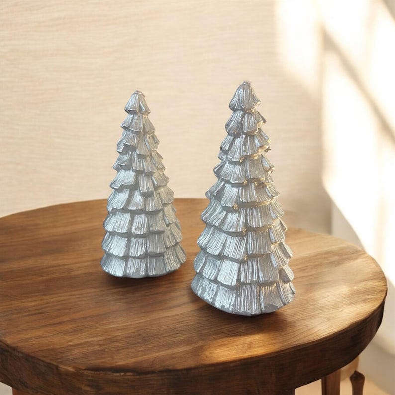 Peut inclure: Deux d&eacute;corations argent&eacute;es en forme de sapins de No&euml;l avec un design textur&eacute; et superpos&eacute;. Elles sont pos&eacute;es sur une table ronde en bois. Les arbres sont de tailles diff&eacute;rentes.