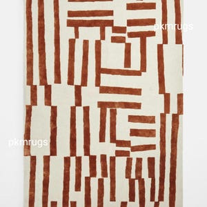 May include: A rectangular rug with a cream-colored background and a repeating pattern of rust-colored vertical and horizontal lines. The lines create a geometric design across the rug's surface. The rug is a decorative floor covering.