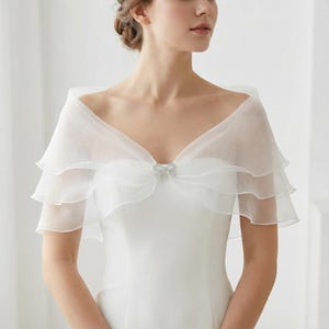 Puede incluir: Un chal de novia blanco y transparente, con capas, con un delicado cierre de pedrería en forma de lazo. El chal presenta un diseño con volantes y se drape sobre los hombros, creando un look elegante y romántico. Ideal para bodas.