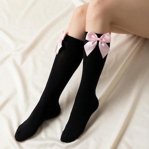 May include: Black knee-high socks with a light pink bow at the top of each sock. The socks are worn on legs, and the background is a white, textured fabric.