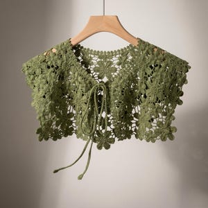 May include: A green crocheted collar with a floral design, hanging on a wooden hanger. The collar features a tie closure at the front and is made of a soft, open-weave material. The color is a muted, olive green.