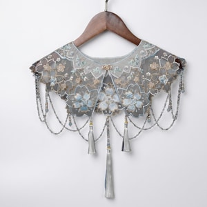 Puede incluir: Un delicado cuello de encaje gris con bordados florales y cadenas de cuentas colgantes. El cuello presenta detalles en azul claro y dorado, con borlas plateadas. Se muestra en una percha de madera sobre un fondo blanco.