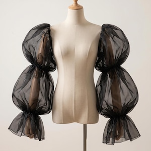 May include: Black sheer puff sleeves displayed on a mannequin. The sleeves have a layered, voluminous design with gathered details at the shoulders, mid-arm, and cuffs. The fabric appears lightweight and airy, creating a dramatic silhouette.