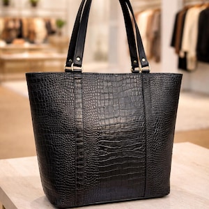Może przedstawiać: Czarna torba typu tote z fakturą imitującą skórę krokodyla. Torba ma dwa czarne uchwyty i złote okucia. Stoi na jasnej powierzchni, z rozmytym tłem wnętrza sklepu.