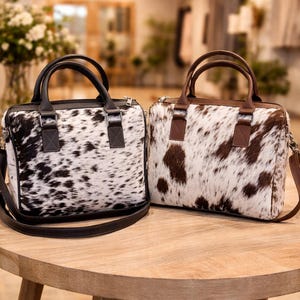 Op de afbeelding: Twee leren handtassen van koeienhuid met bruine handvatten en riemen staan op een houten tafel. De ene tas heeft zwart-witte vlekken, de andere bruin-witte vlekken. Beide hebben een rechthoekige vorm en een ritssluiting.