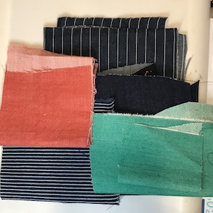 以下が含まれることがあります： 様々な色と柄の生地見本。無地の赤、青と白のストライプ、無地の緑、黒と白のストライプなどがあります。
