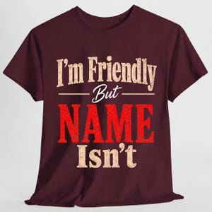 May include: A maroon short-sleeve t-shirt featuring the text "I'm Friendly But NAME Isn't" printed on the front. The words "I'm Friendly" and "Isn't" are in a distressed beige font, while the word "NAME" is in a large, bold, distressed red font.