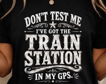 May include: A black short-sleeved t-shirt featuring white distressed-style text that reads: DON'T TEST ME I'VE GOT THE TRAIN STATION IN MY GPS. The design includes decorative flourishes, stars, and curved lines surrounding the bold, capitalized lettering.